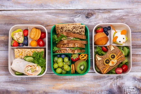 Three food containers on a wooden table: one with salad, one with pasta, and one with soup.